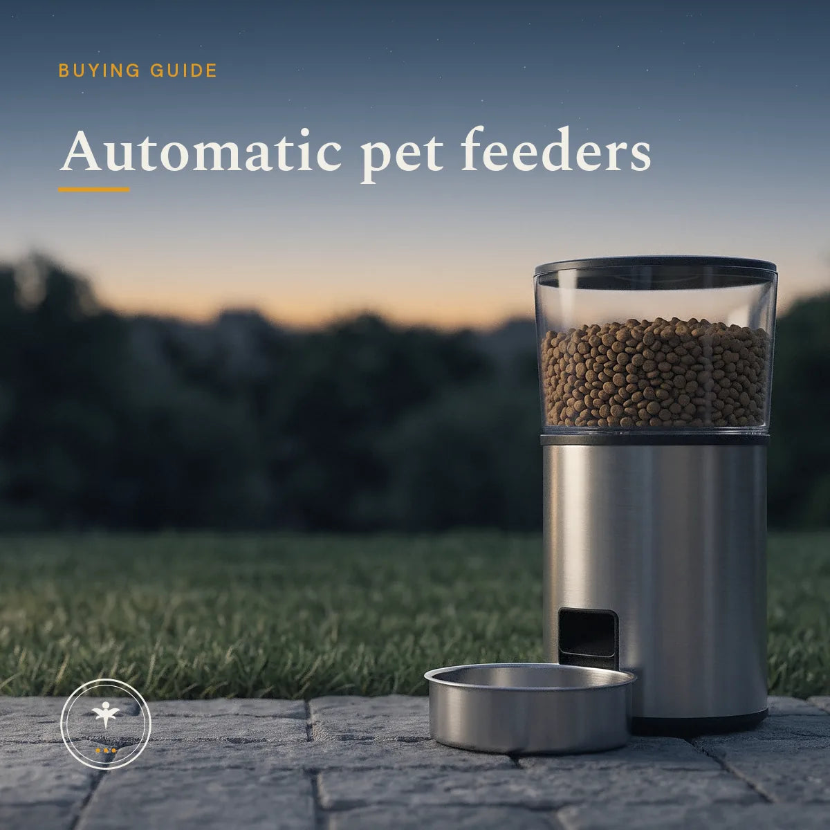 A plain stainless automatic pet feeder with dry food in its clear hopper, beside a stainless bowl on stone paving at the edge of a lawn at dusk, with a soft dark treeline behind.